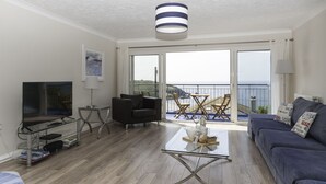 Una sala moderna con televisor, asientos, una mesa de centro y vista al mar a través de amplios ventanales.