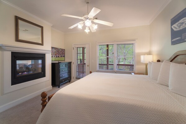 A bedroom with a fireplace, a ceiling fan, a large bed, and a view of the outdoors through a door.