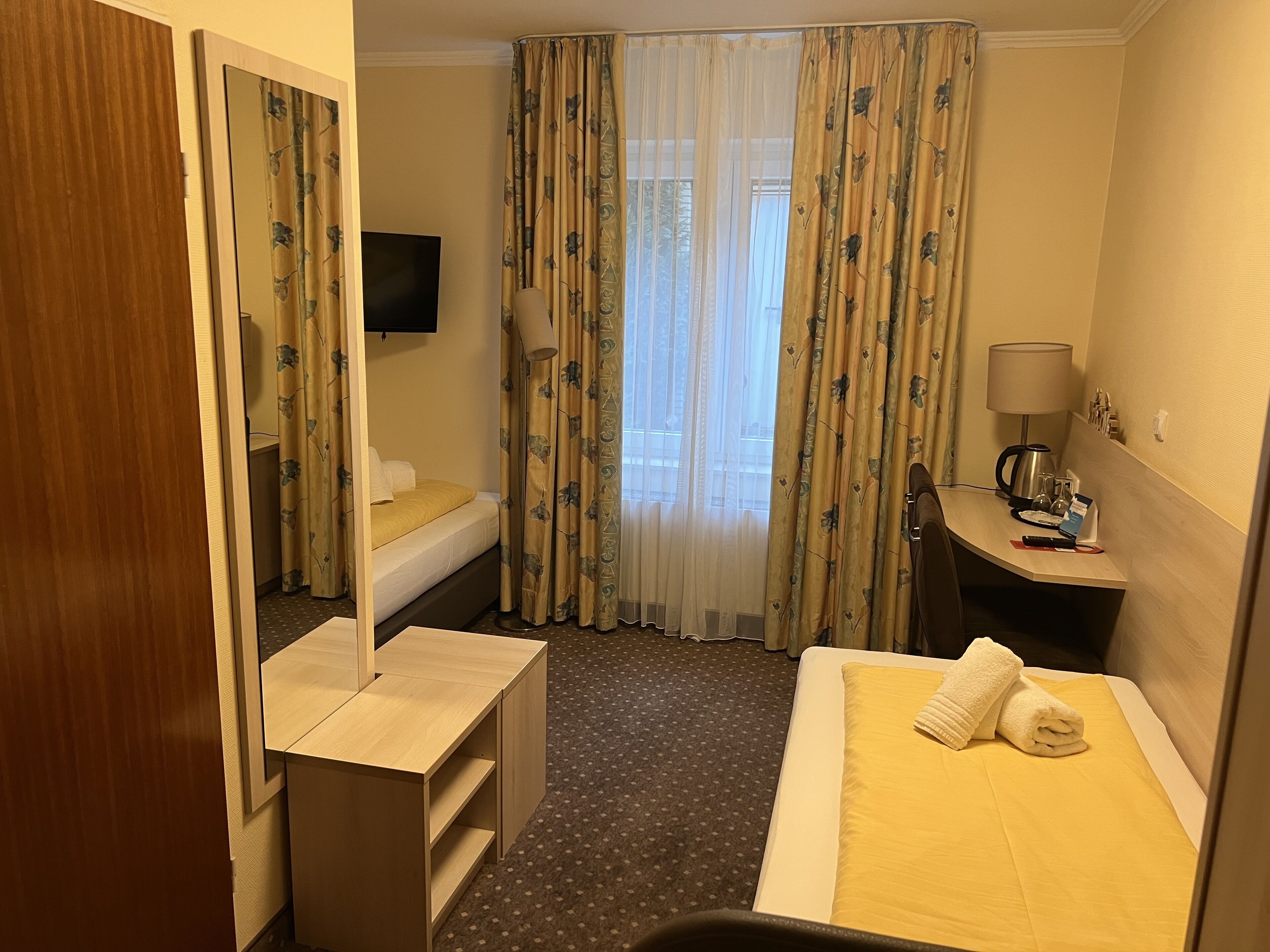 A hotel room with a bed, a desk, a chair, a TV, and a window with curtains.