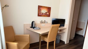 Ein kleines, möbliertes Zimmer mit Schreibtisch, zwei Stühlen, einem Fernseher, einem Gemälde und einem Mülleimer.