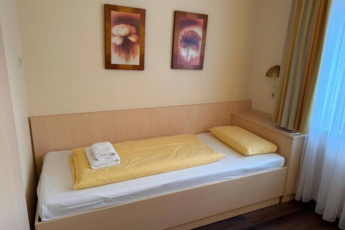 Un lit simple avec une couvre-lit jaune et un oreiller blanc, une serviette pliée, et deux tableaux représentant des motifs floraux, encadrés et accrochés au mur.