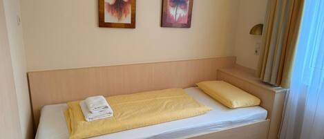 A single bed with a yellow bedspread and a white pillow, a folded towel, and two framed floral paintings on the wall.