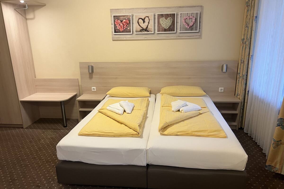 Une chambre d’hôtel avec deux lits, un petit bureau et un cadre photo fixé au mur, orné de décorations en forme de cœur.