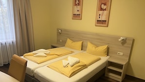 Ein Hotelzimmer mit zwei Betten, jeweils mit gelber Bettwäsche und weißen Handtüchern. Es gibt einen hölzernen Kopfteil, einen Nachttisch und einen Stuhl. Der Raum hat einen Holzboden und ein Fenster mit Vorhängen.