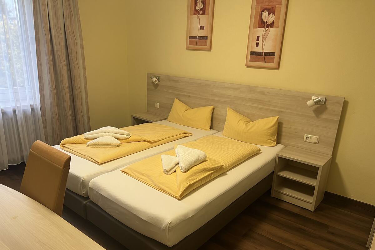 Une chambre d’hôtel avec deux lits, chacun agrémenté de draps jaunes et de serviettes blanches. On y trouve une tête de lit en bois, une table de chevet et une chaise. La chambre est dotée d’un plancher en bois et d’une fenêtre avec des rideaux.