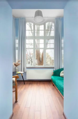 Una habitación con un ventanal grande, un radiador, un sofá verde y una mesita con un florero.
