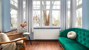 Um quarto com um sofá verde, uma cadeira de madeira, uma pequena mesa com um vaso e uma janela grande com cortinas.