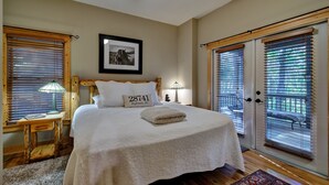 A bedroom with a large bed, wooden headboard, two bedside tables with lamps, a framed picture on the wall, and a view through the window blinds.
