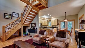 A spacious living area with a wooden staircase, leather sofas, a dining table, and a kitchen in the background.