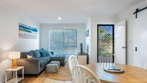 A modern living room with a grey sofa, a dining table with chairs, a TV, and a window with blinds. - The Weekender - spacious townhouse in central location (Evans Head)