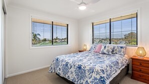 A bedroom with a bed, two bedside tables, a ceiling fan, and windows with blinds.