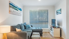 A modern living room with a grey sofa, a TV stand, and a large window with blinds. - The Weekender - 4-46 Cypress Street (Evans Head)