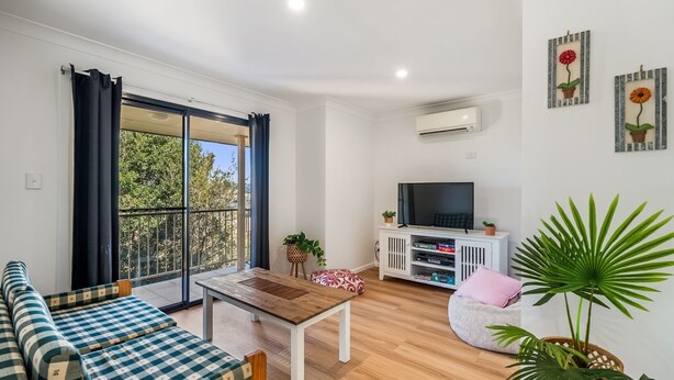 A living room with a sofa, a coffee table, a TV on a stand, and a large window with curtains. - The Weekender - spacious townhouse in central location (Evans Head)