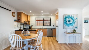 A modern kitchen with wooden cabinets, a dining table with white chairs, a stainless steel dishwasher, and a large abstract painting on the wall. - The Weekender - spacious townhouse in central location (Evans Head)