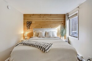 Un dormitorio con cama, cabecera de madera, mesita de noche, lámpara y ventana con vistas.