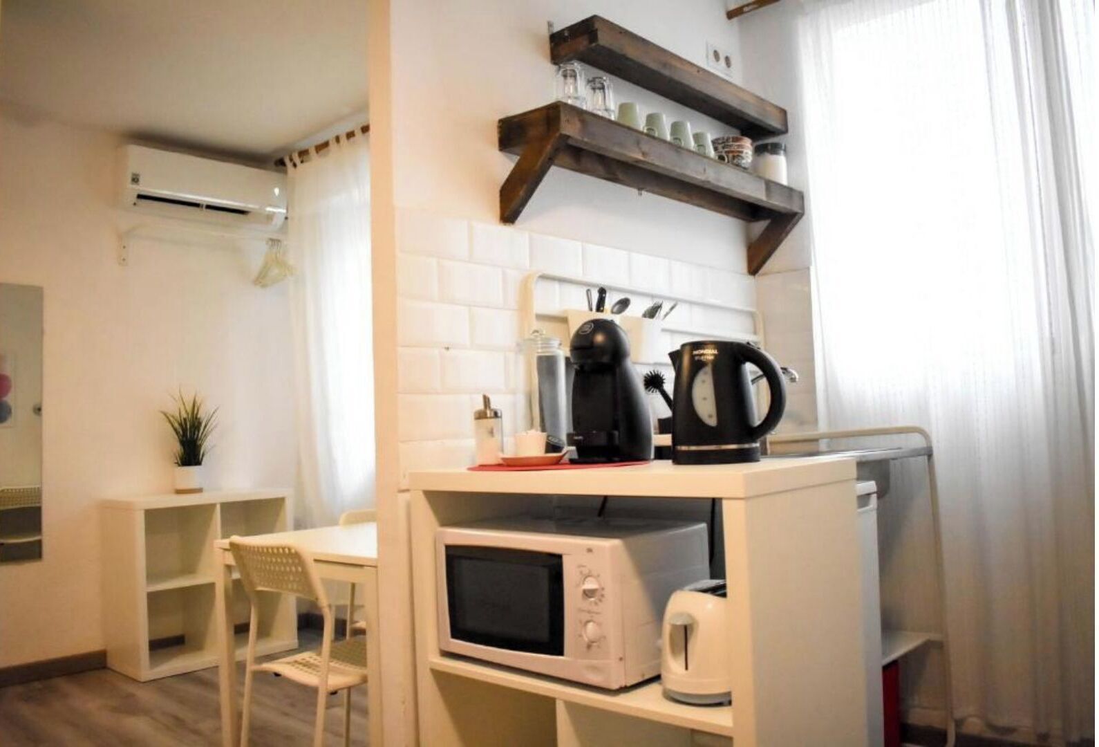 Une petite cuisine équipée d’un four à micro-ondes, d’un grille-pain et d’une machine à café, le tout posé sur un plan de travail blanc. On y trouve également une chaise et une petite étagère contenant des ustensiles de cuisine.