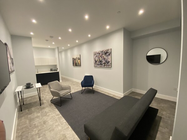 Una sala moderna con un sofá gris, un sillón azul y un sillón gris con patas metálicas. Hay una mesita blanca con un cuenco y una taza. La habitación tiene una alfombra gris y paredes blancas.