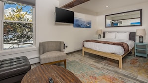 A modern hotel room with a bed, a television, a chair, a coffee table, and a window with a view of trees and mountains. - Newly remodeled studio w/gorgeous mountain views near Big Sky Resort - Hill Condo Lookout (Big Sky)
