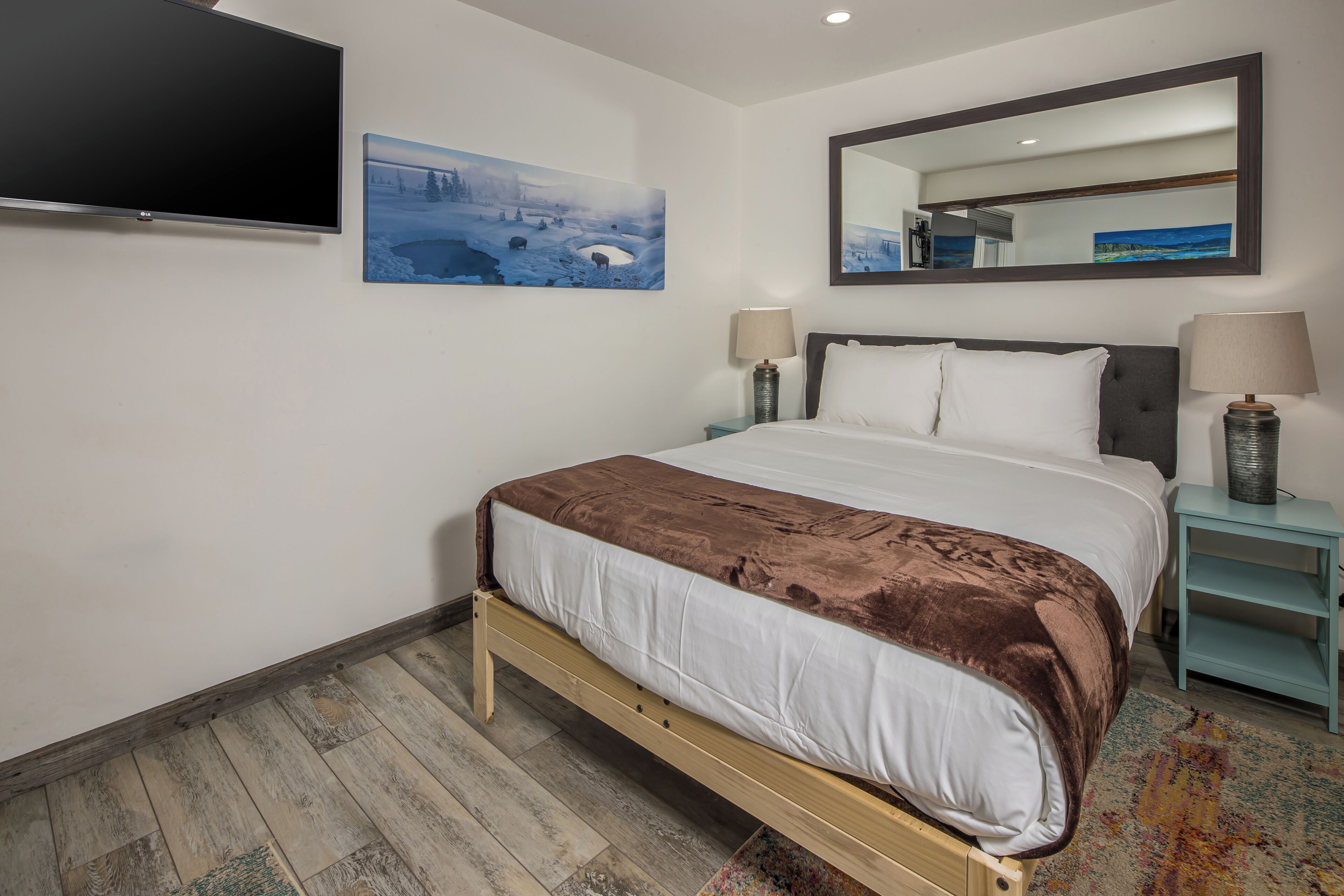 A bedroom with a bed, a TV, a painting, a mirror, and a nightstand with a lamp.