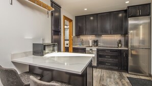 A modern kitchen with dark cabinets, a stainless steel refrigerator, a microwave, and a bar area with two chairs. - Newly remodeled studio w/gorgeous mountain views near Big Sky Resort - Hill Condo Lookout (Big Sky)