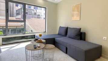 Uma sala de estar moderna com um sofá modular cinzento, duas mesas redondas e uma janela grande com vista para prédios.