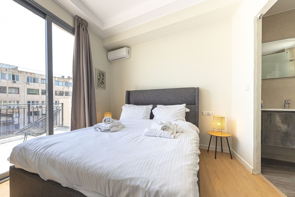 A hotel room with a large bed, a bedside table with a lamp, a bathroom with a mirror, and a view of buildings outside.