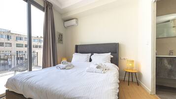 Quarto de hotel com uma cama grande, mesa de cabeceira com abajur, banheiro com espelho e vista para edifícios.