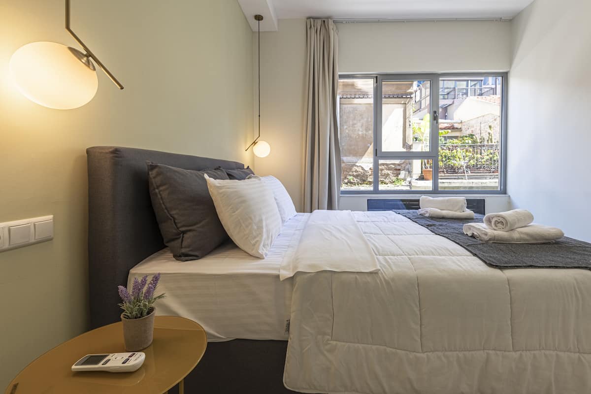 A hotel room with a bed, a bedside table with a lamp, a window with curtains, and a view of the outside.
