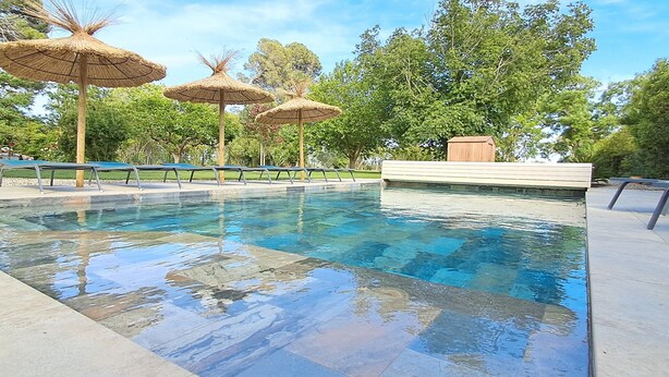 Ein Schwimmbecken mit klarem Wasser, umgeben von einer gefliesten Terrasse und beschattet durch Strohdachschirme.