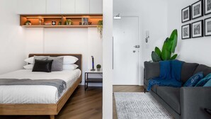 A modern bedroom with a bed, bedside table, and a shelf with books and plants. The room has a wooden floor and white walls. - Stylish Studio Near Kings Cross Train Station (Elizabeth Bay)