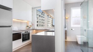 A modern kitchen with white cabinetry, a built-in oven, and a large window overlooking a residential building. - Stylish Studio Near Kings Cross Train Station (Elizabeth Bay)