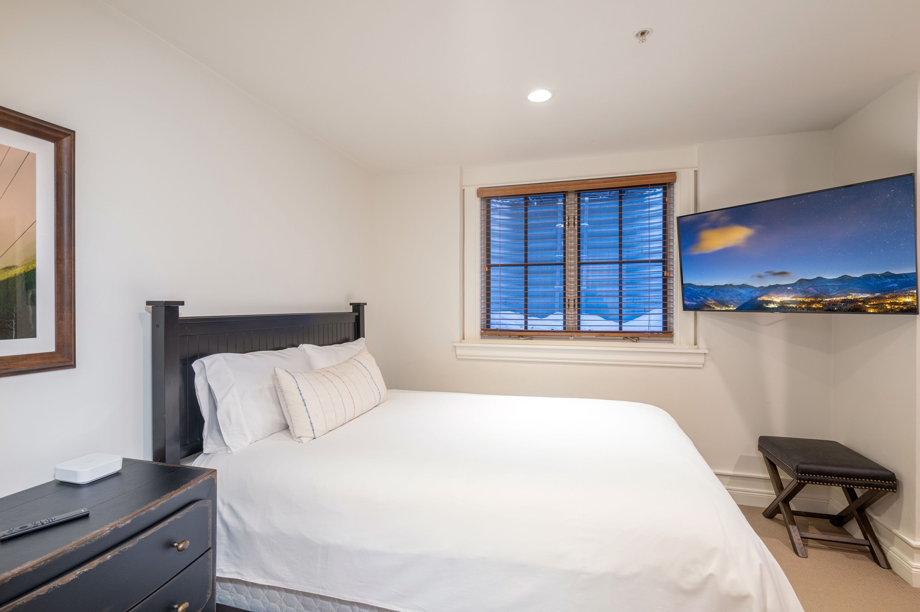 A bedroom with a bed, a nightstand, a window with blinds, and a painting on the wall.
