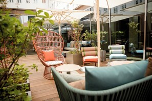 A patio area with wicker chairs, a round table, and a cushioned sofa. There are potted plants and a large umbrella providing shade. - Motel One Hannover-Oper (Hannover)