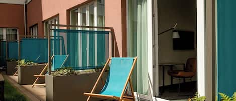 Un balcon avec deux chaises en bois et un paravent en tissu bleu, une petite table, et une chaise à l’intérieur d’une pièce où se trouve un téléviseur.