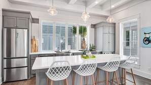 Une cuisine moderne dotée d’appareils électroménagers en acier inoxydable, d’un îlot central blanc avec des tabourets de bar et de grandes fenêtres offrant une vue sur l’extérieur.
