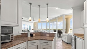 Una cocina moderna con armarios blancos, encimeras de granito y una isla integrada. Hay luces colgantes, un ventilador de techo y vistas al exterior a través de amplias ventanas.
