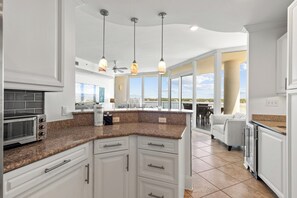 Una cucina moderna con mobili bianchi, piani di lavoro in granito e un'isola integrata. Sono presenti luci a sospensione, una ventola a soffitto e vista all'esterno attraverso ampie finestre.