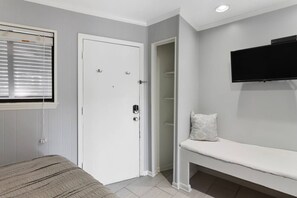 A room with a bed, a white door, a window with blinds, a white bench, and a flat-screen TV mounted on the wall.