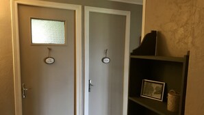 Un couloir avec deux portes, chacune étant munie d’une plaque nominative et d’une petite fenêtre. Une étagère, sur laquelle se trouvent un tableau encadré et un panier, est placée sur le côté droit.