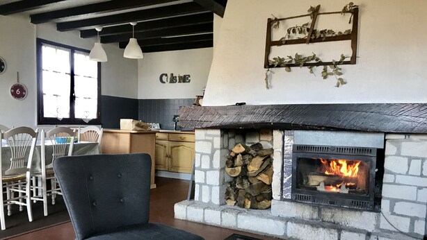 Un salon chaleureux avec une cheminée en pierre, un coin repas avec des chaises en bois et une cuisine à l’arrière-plan.