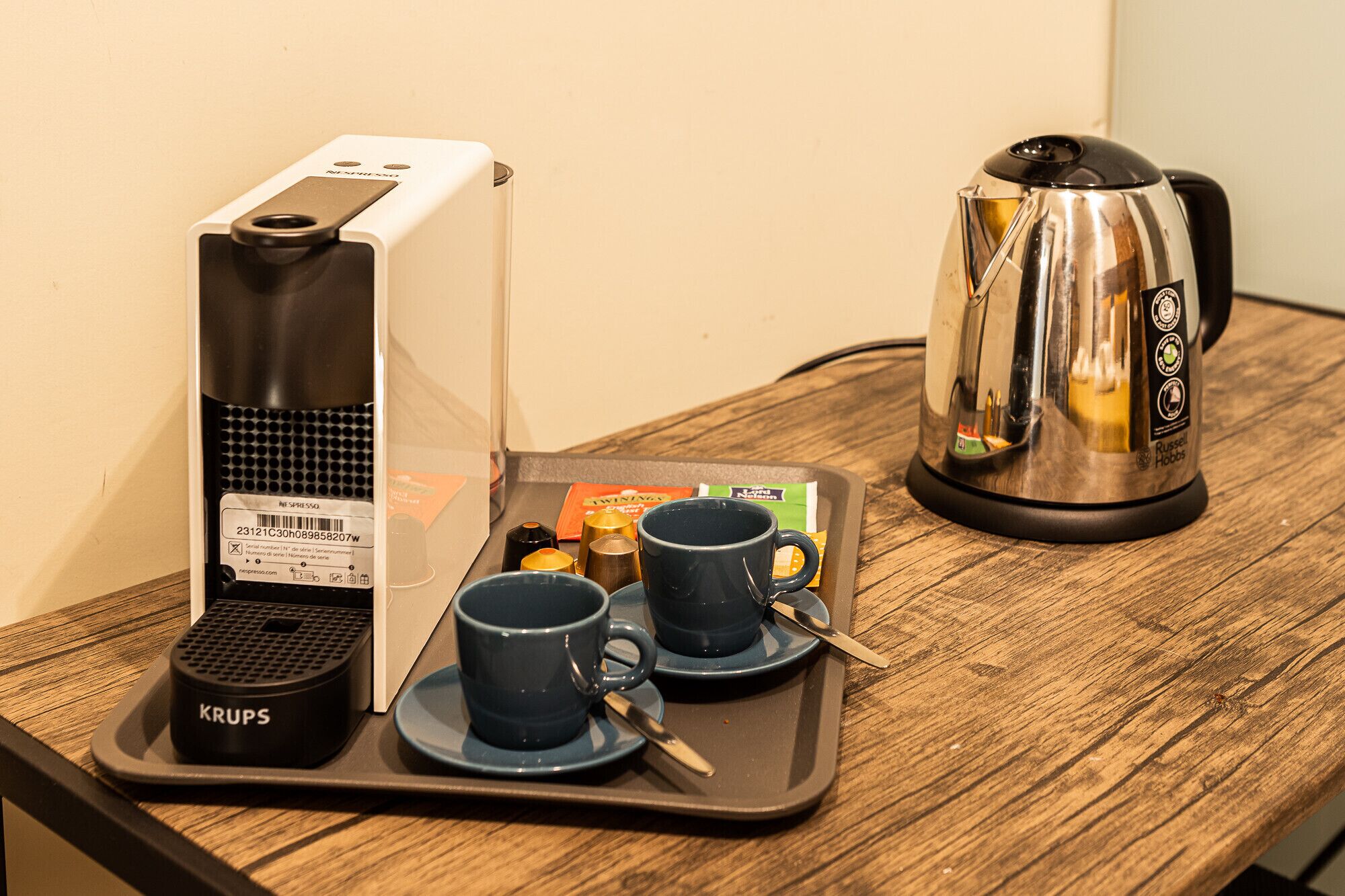 Una cafetera, un hervidor y una bandeja con tazas y acompañamientos sobre una mesa de madera.