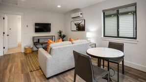 A modern living room with a grey sofa, orange pillows, a flat-screen TV, a mounted air conditioner, a dining table with chairs, and a window with blinds.