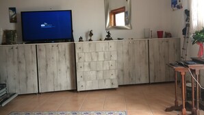 Un salon avec un téléviseur affichant un symbole Wi-Fi, un meuble en bois, une petite table avec une plante et un sol carrelé.