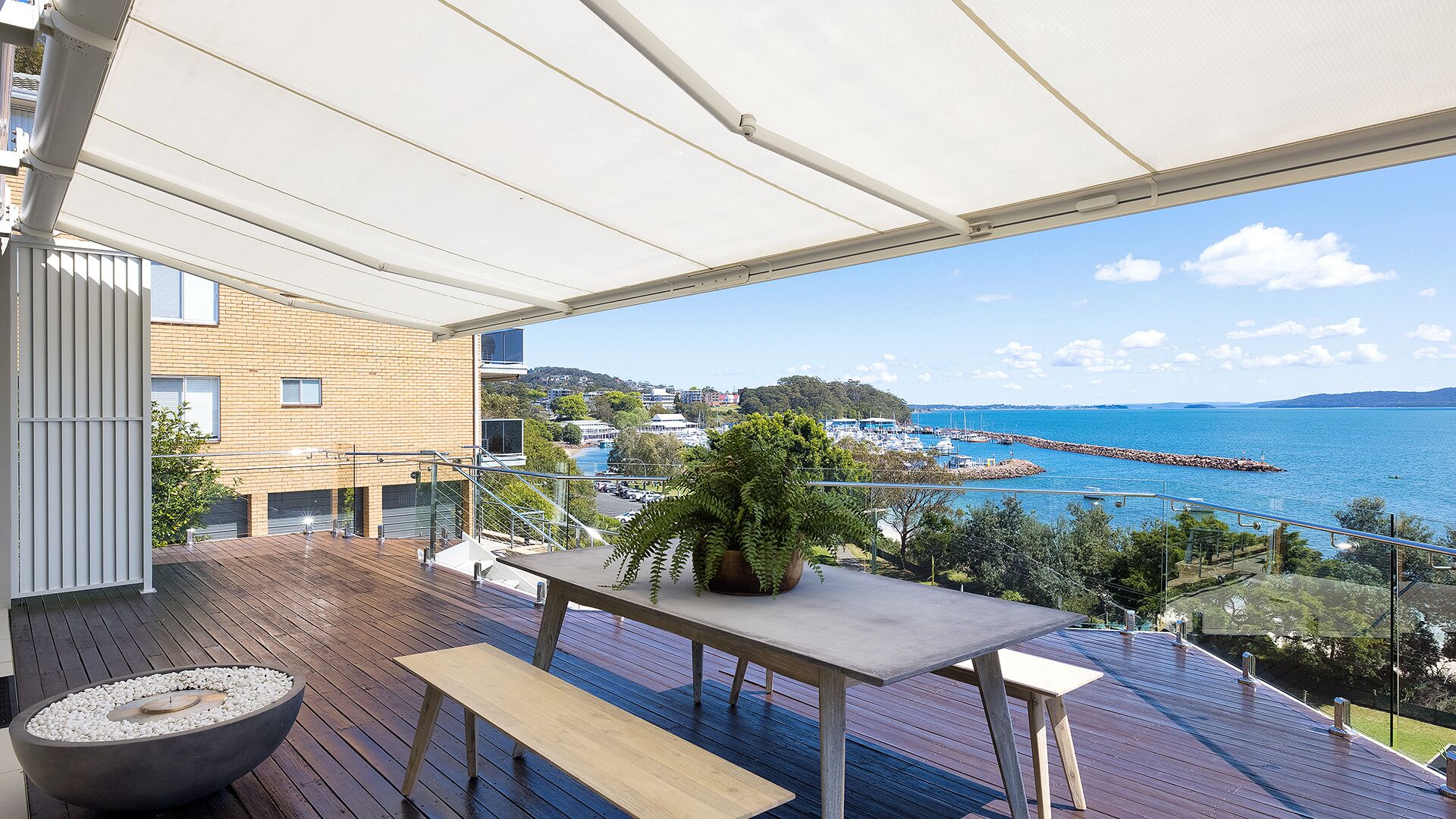 A modern balcony with a view of the sea, wooden flooring, a table, and a potted plant.