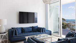 Uma sala de estar moderna com uma grande televisão de tela plana, um sofá azul, uma mesa de centro branca e vista para uma marina através de uma porta de vidro.