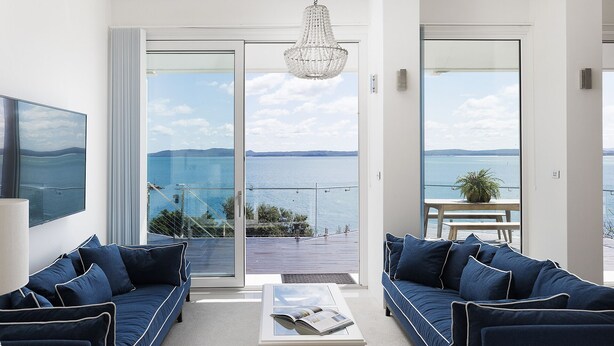 Uma sala de estar moderna com uma janela ampla que oferece vista para o mar e terra distante. Há um sofá, uma mesa de centro e um lustre.