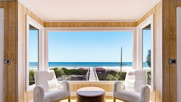 A room with a large window overlooking the beach, two white armchairs, and a wooden ottoman.