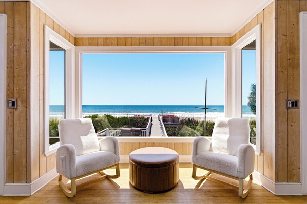 A room with a large window overlooking the beach, two white armchairs, and a wooden ottoman.