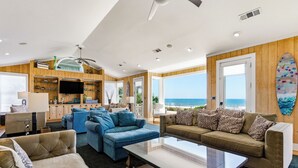 A spacious living room with a large window overlooking the ocean, a surfboard on the wall, and a television mounted above a wooden cabinet.