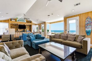 A spacious living room with a large window overlooking the ocean, a surfboard on the wall, and a television mounted above a wooden cabinet.